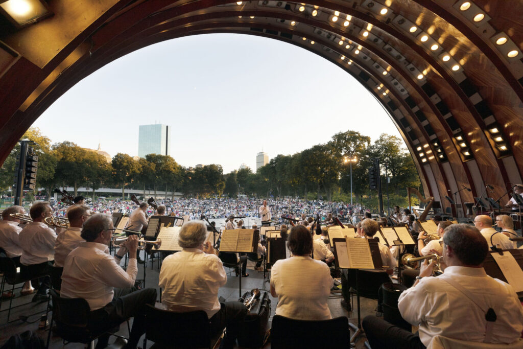 Symphony orchestra performing in an open-air bandshell before a large crowd.