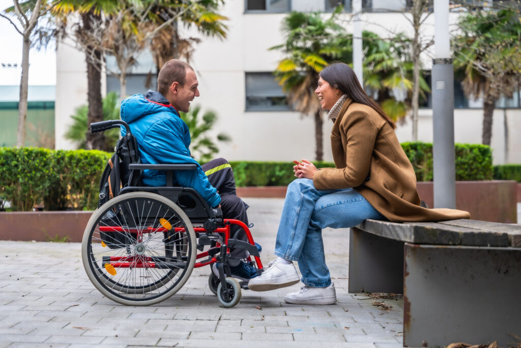 Man with a disability in a wheelchair having a cheerful conversation with a woman sitting on a park bench, sharing a moment of connection and inclusion outdoors