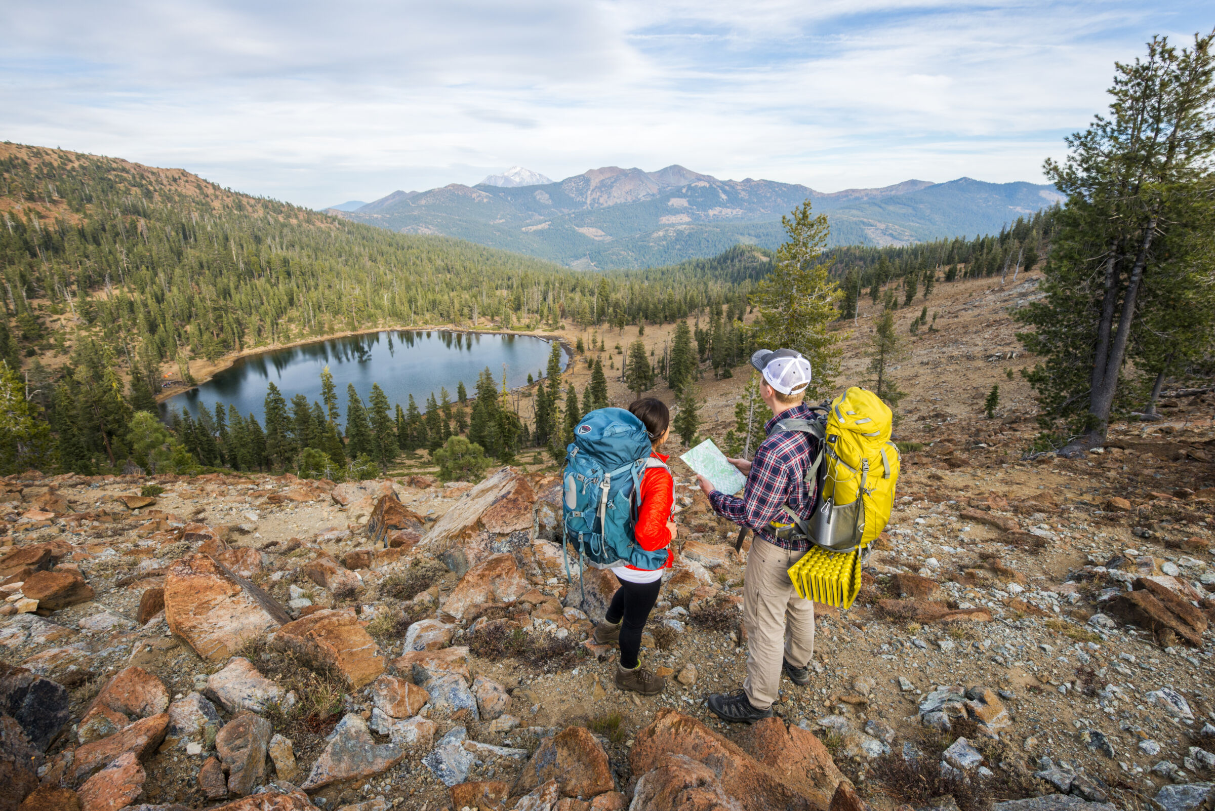 Two hikers overlooking a mountain lake and forested landscape while reviewing a trail map.