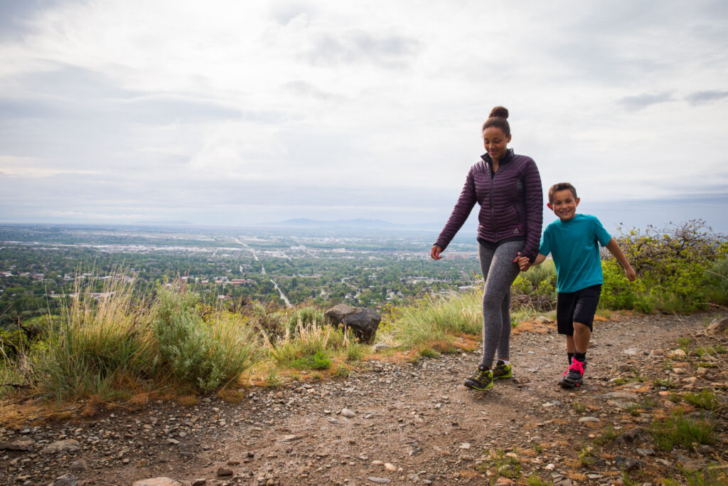 Adult and child hiking on a trail overlooking a city and surrounding landscape.